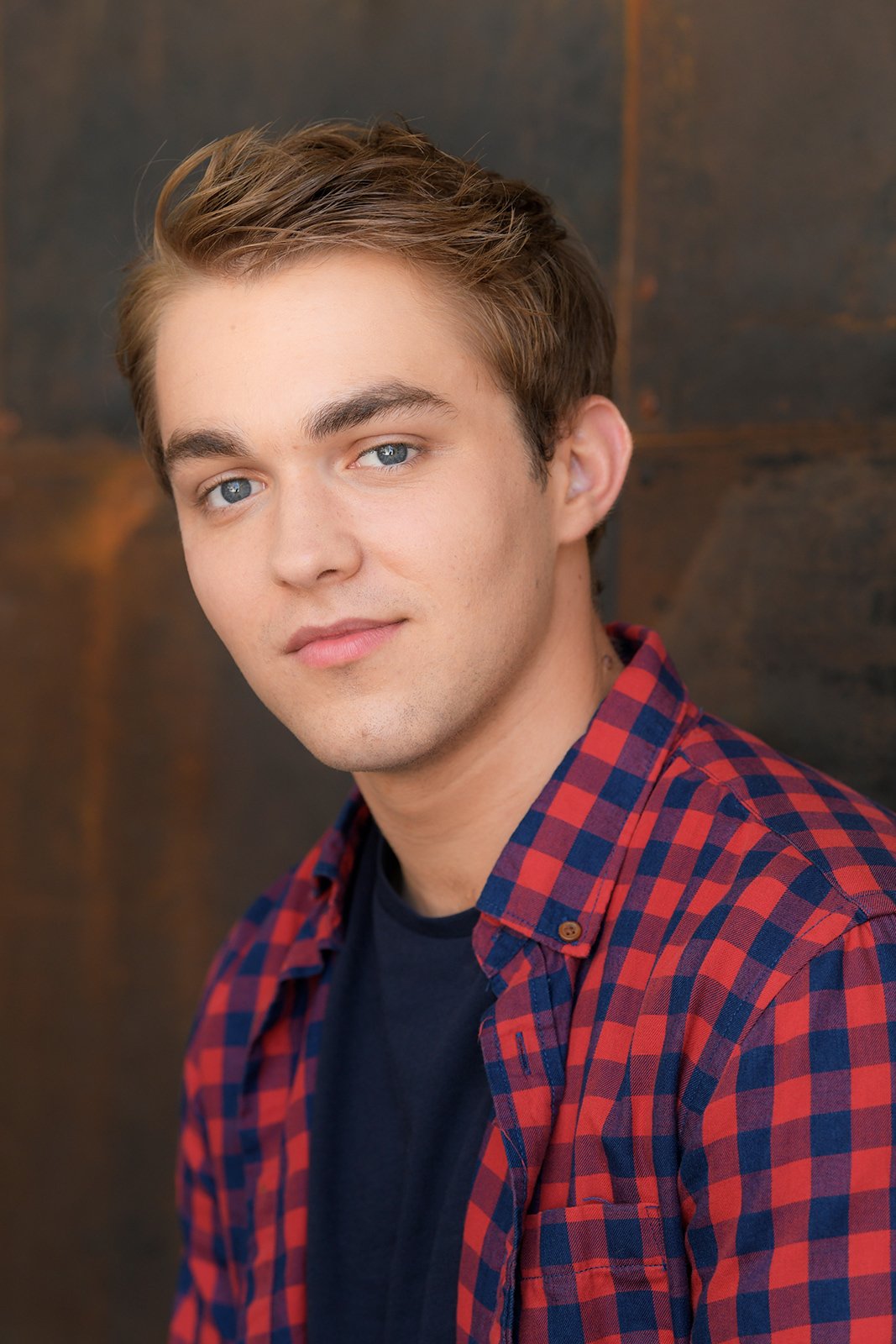 A young man with light brown hair and blue eyes wearing a red and blue plaid shirt over a black t-shirt, smiling softly, with a blurred dark wooden background.